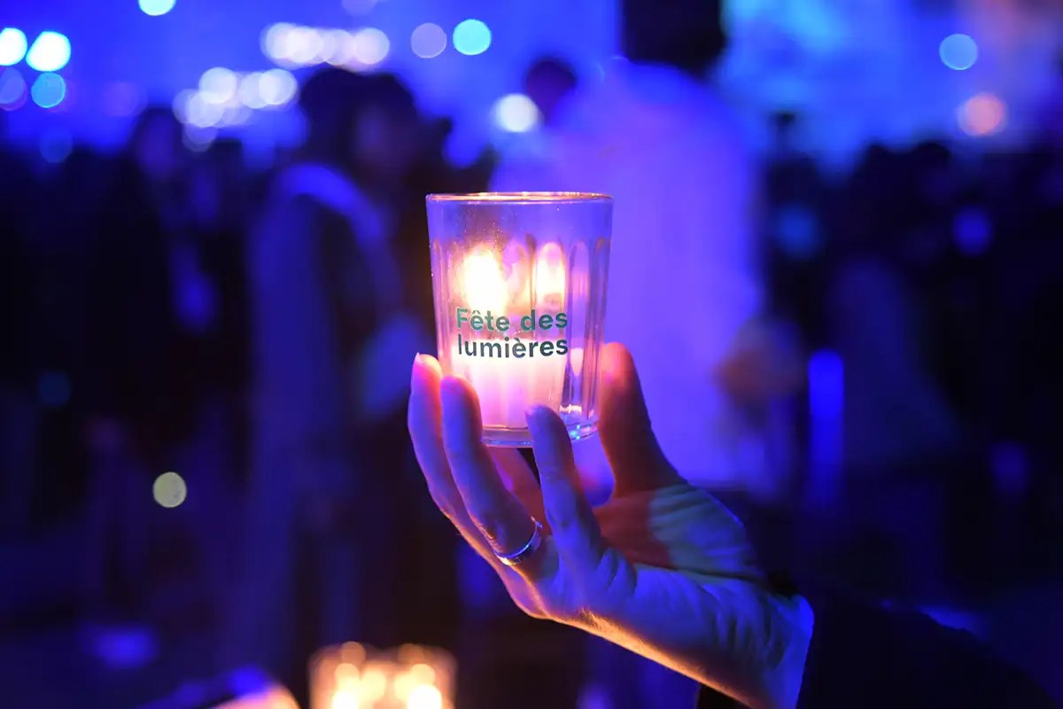 Une femme tient un verre marqué « Fête des lumières » contenant une bougie , devant une foule floue baignée de lumière bleue.
